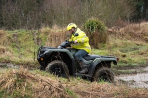 Man in high vis and helmet driving an ATV up an incline in a wooded area
