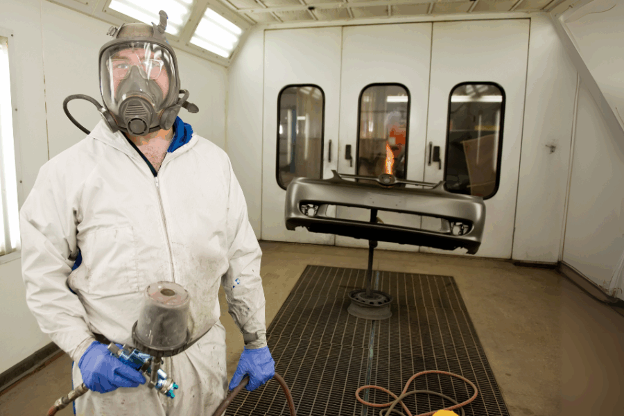 A paint strayer wearing PPE including a air-fed respirator, standing in a spray booth