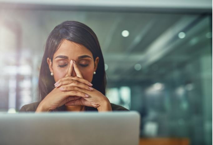 A woman sitting at a desk with her eyes closed and hands resting against her face while working on a laptop, suggesting she may be feeling overwhelmed or stressed at work.